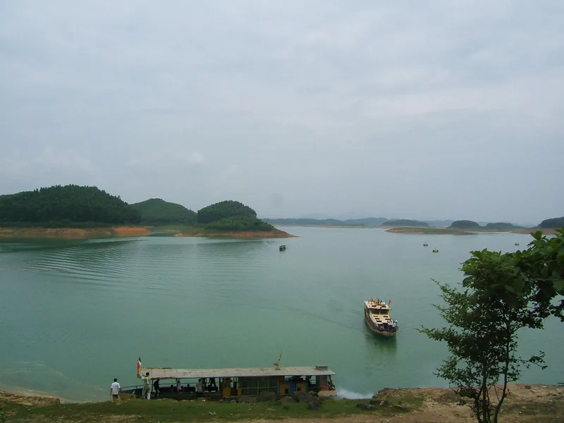 Một chiếc thuyền gỗ băng qua mặt nước xanh yên bình của hồ Thác Bà, điểm xuyết những đảo nhỏ phủ rừng.