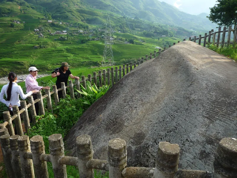 Du khách đứng bên bãi đá cổ có khắc hình được rào chắn, nhìn xuống ruộng bậc thang xanh ở Sapa.